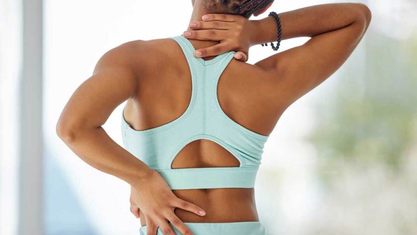 A brown skinned woman in workout wear next to a window, holding her back & neck due to pain and discomfort.