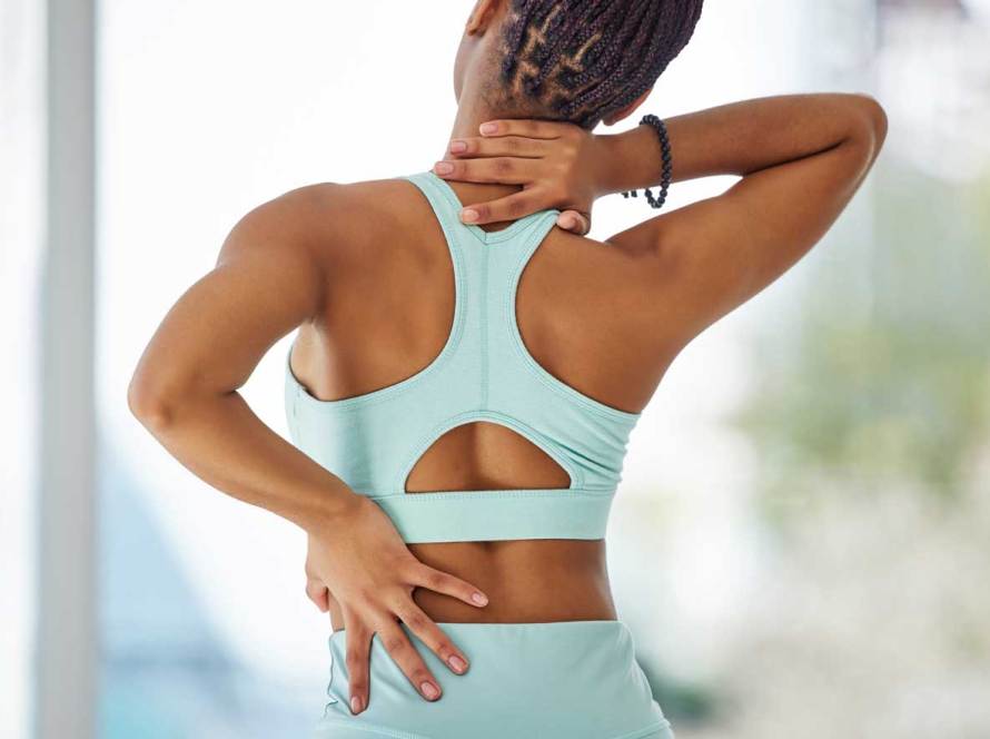A brown skinned woman in workout wear next to a window, holding her back & neck due to pain and discomfort.