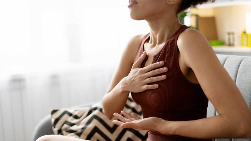a light brown skinned woman sits on the couch while dramatizing the flow of breathe in & out of the body using breathing exercises presented in the blog post.